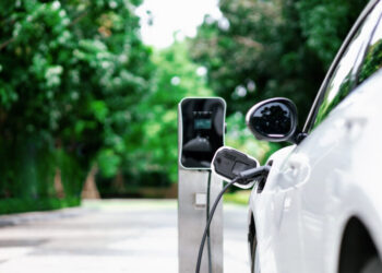 Focus closeup electric vehicle plugged in with EV charger device from blurred background of public charging station powered by renewable clean energy for progressive eco-friendly car concept.